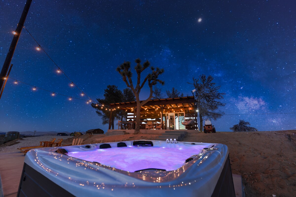 A spacious hot spa glows with soft purple lighting, set against a backdrop of a serene night sky filled with stars. The contemporary house features bright outdoor lighting and a relaxed seating area, enhancing the tranquil desert ambiance.