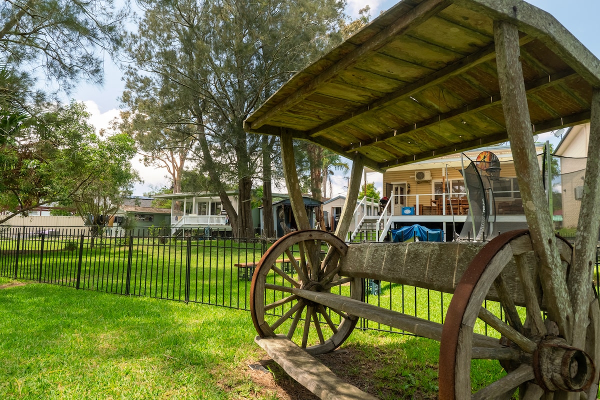 A rustic wooden cart is positioned in the foreground, surrounded by a lush green lawn. Behind it, the vintage-style cottage and a spacious deck are visible, creating a welcoming outdoor space. A black fence encloses the yard, enhancing the peaceful atmosphere.