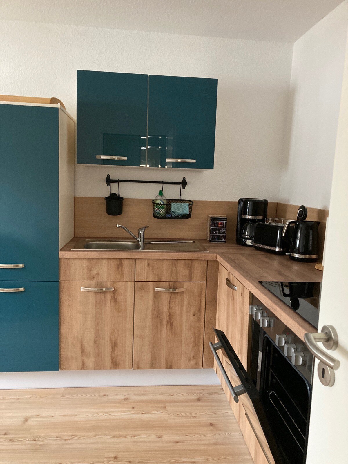 A compact kitchen area features wooden cabinetry paired with teal upper cabinets. A stainless-steel sink sits under the mounted shelf, which holds essential items. Appliances include a coffee maker, kettle, and a stovetop oven, maintaining a tidy and functional aesthetic.
