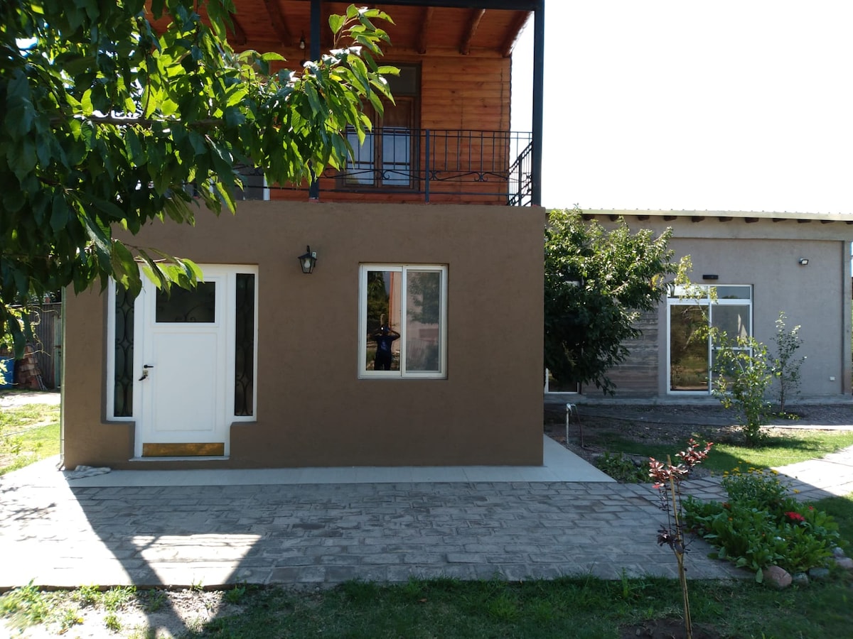 The exterior of a two-story house is shown, featuring a warm brown facade and a balcony. A front door is visible, along with several windows that allow natural light inside. A garden area with greenery and paved pathways complements the inviting atmosphere.