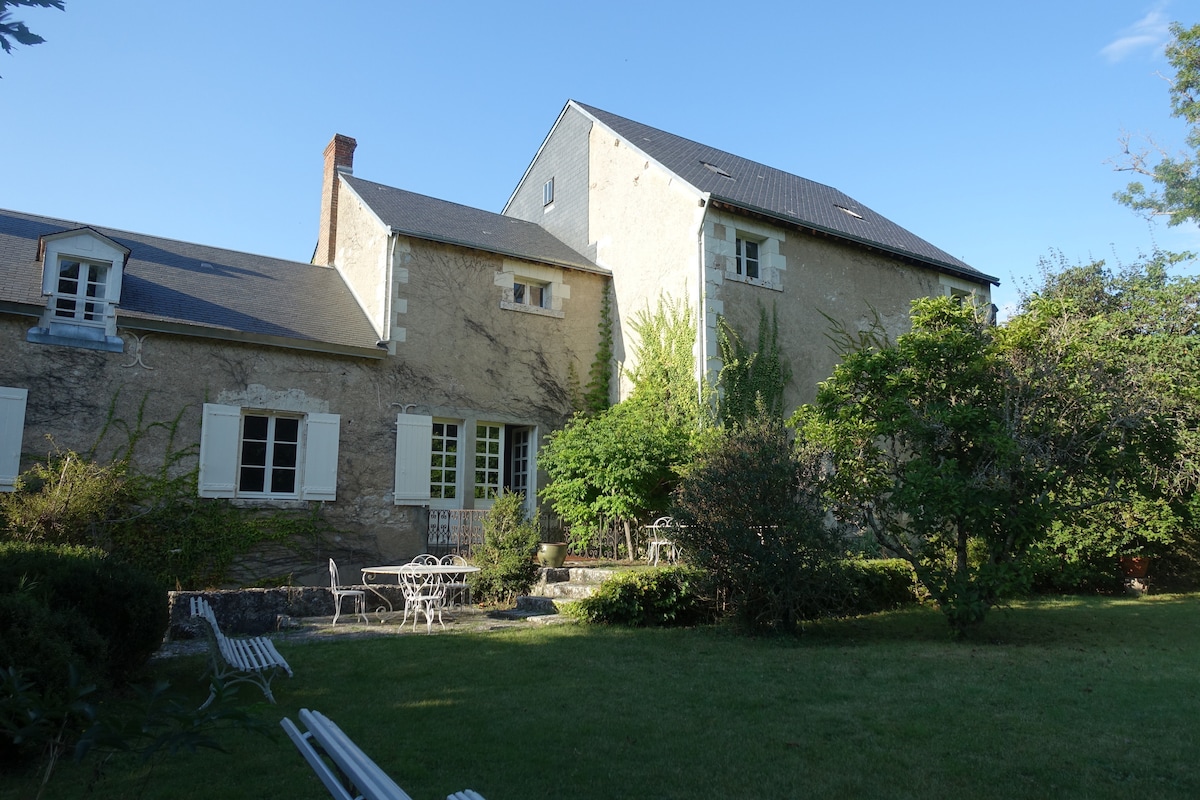 The exterior of the house reveals a rustic façade with white windows that frame the structure. A well-maintained garden features a small seating area with a round table and chairs, surrounded by greenery. A clear blue sky adds to the serene atmosphere of the setting.