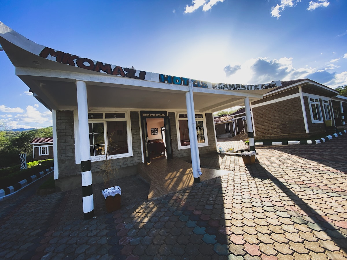 The entrance of Mkomazi Hotels is characterized by a spacious covered porch, with a sign prominently displaying the hotel's name. The surrounding area is paved with colorful stones, and the structure features large windows, allowing ample natural light into the interior.