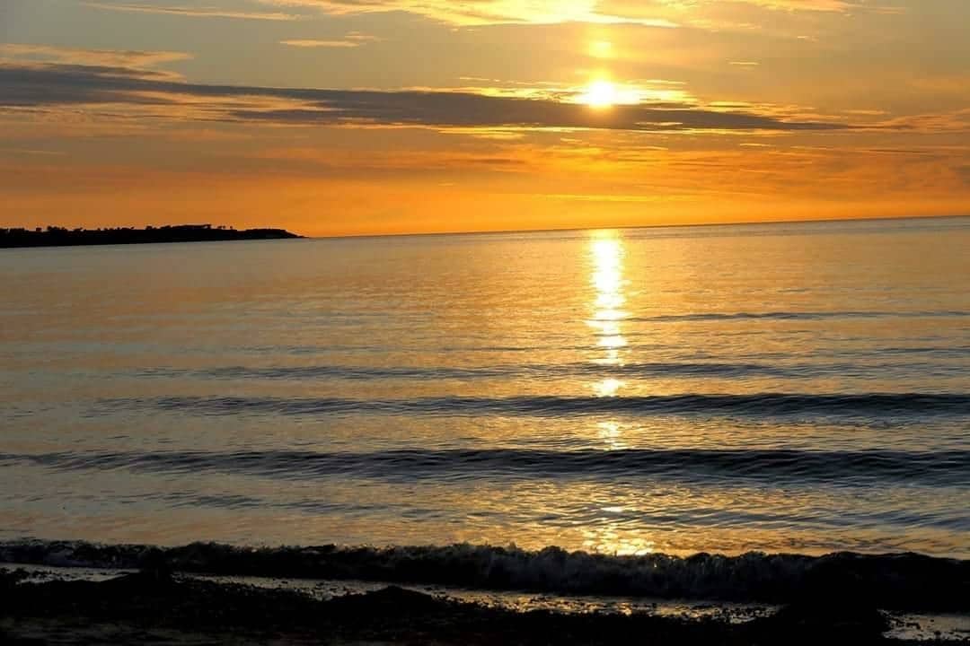 A serene sunset reflects off calm waters, creating a warm golden hue across the horizon. Gentle waves ripple towards the shore, framed by a subtle shoreline. The distant coastline is visible against the evening sky, enhancing the peaceful atmosphere of the beach.