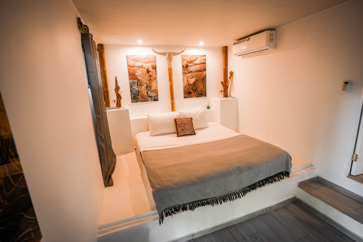 A tranquil sleeping area features a spacious bed with light-colored linens and a cozy gray throw. Two artistic pieces decorate the walls, complemented by sculptures on either side. An air conditioning unit is mounted above, providing comfort in the serene room.