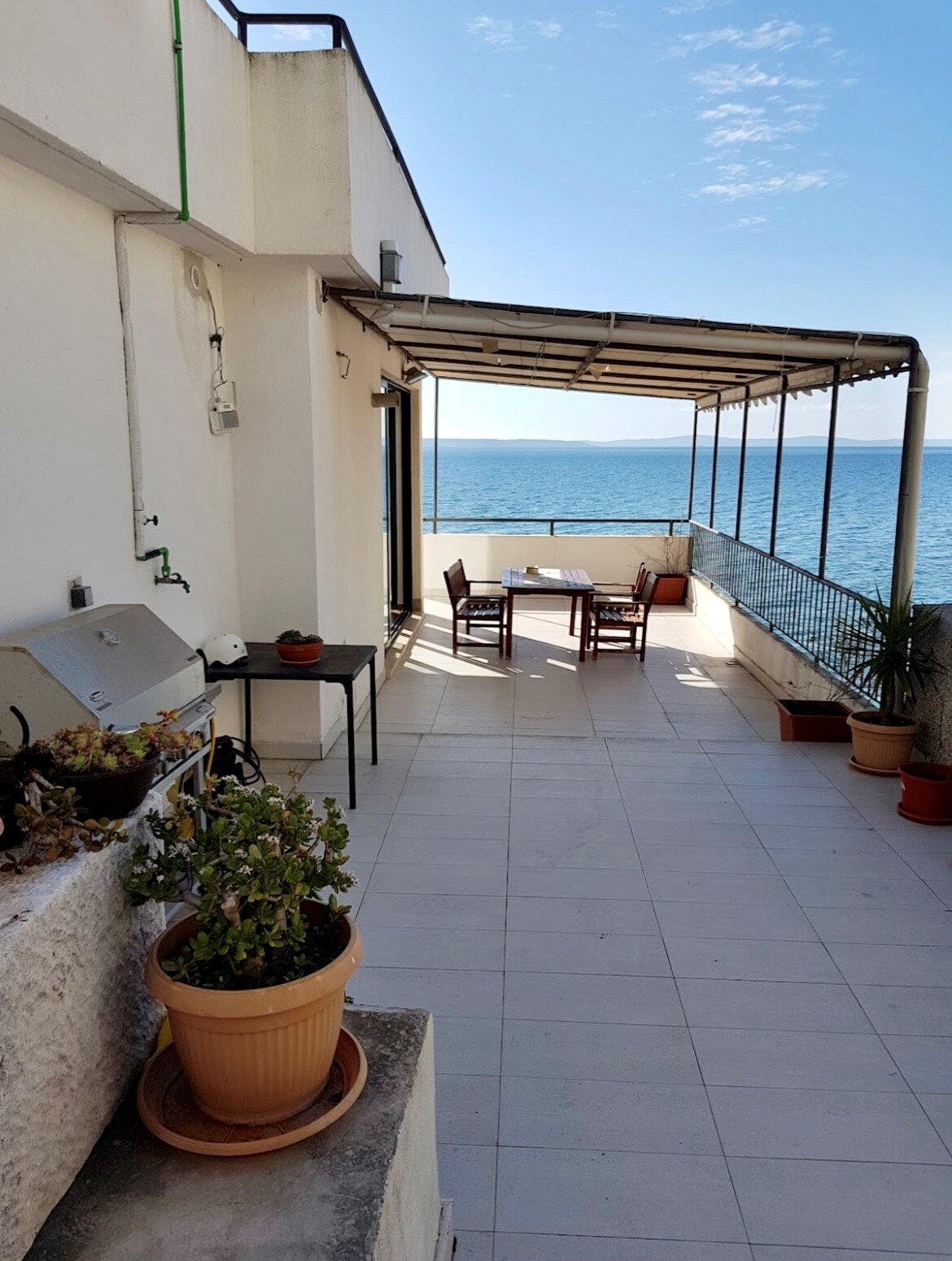 An outdoor terrace features a dining set with wooden chairs and a table, overlooking the expansive ocean. Potted plants add greenery to the space, while a BBQ grill is positioned against the wall. The serene view of the turquoise sea enhances the outdoor experience.