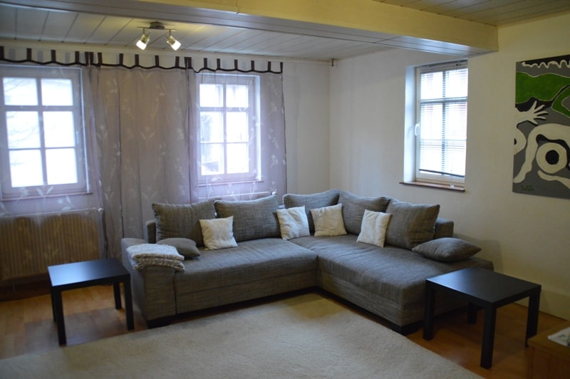 A spacious living room features a large sectional sofa adorned with decorative pillows. Two small tables sit beside the sofa, complementing the light wood flooring. Natural light enters through two windows, each dressed with sheer curtains, creating a relaxed atmosphere.