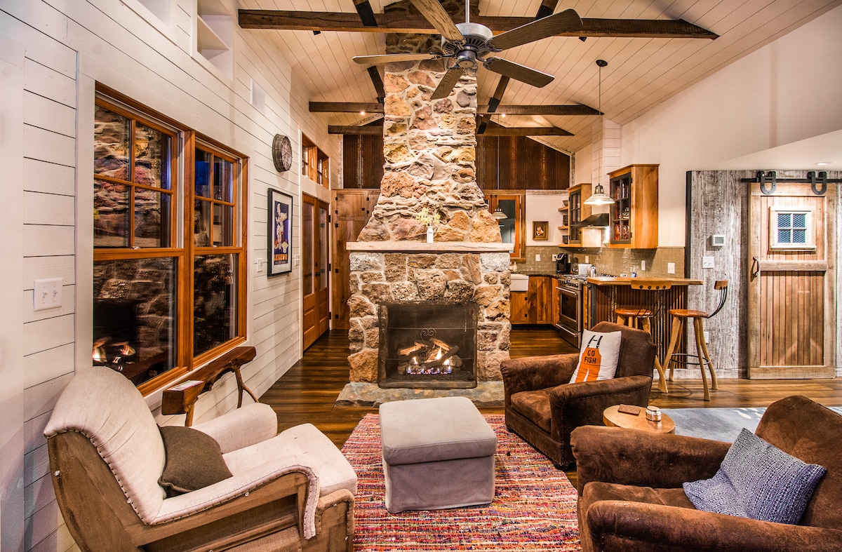 A cozy living room features a stone fireplace and wooden accents. Two plush armchairs and a soft ottoman are arranged around a colorful area rug. Large windows allow for ample natural light, while the open layout connects to the kitchen in the background.
