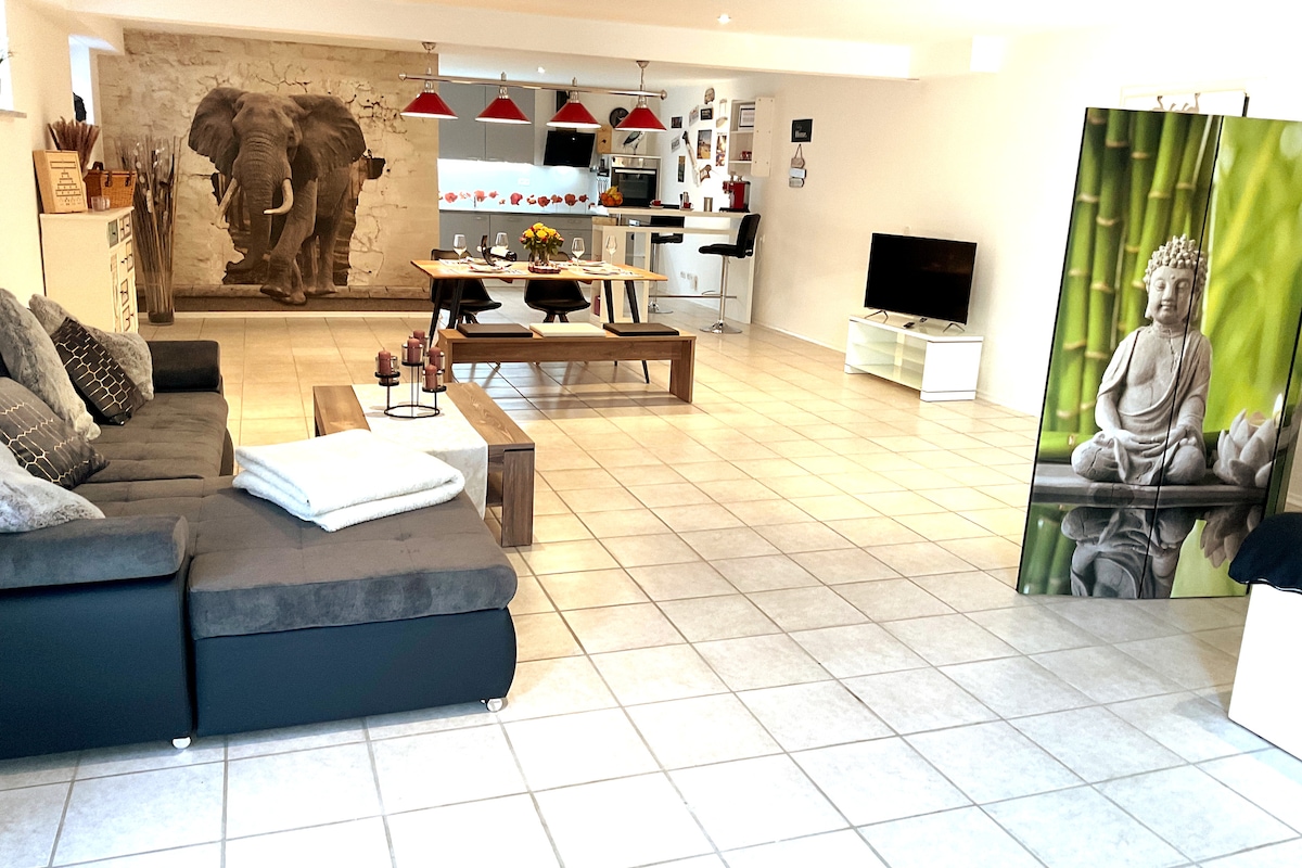 The open-plan living area features a comfortable gray sofa with decorative cushions. A large dining table with rustic charm is positioned centrally, surrounded by a modern kitchen with red pendant lights above. A tv stands against a white wall, while artful accents, including a bamboo and Buddha decor, enhance the space.