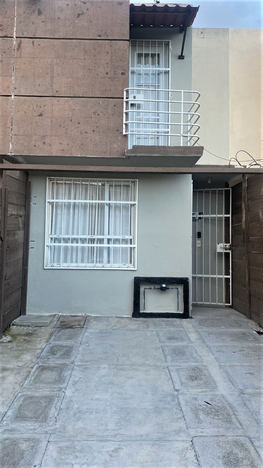 The entrance to the property is showcased, featuring a clean, paved path leading to a secure metal gate. A window with grilles is visible on the right, while the left side displays a small balcony above the entryway. The overall design reflects a modern aesthetic.