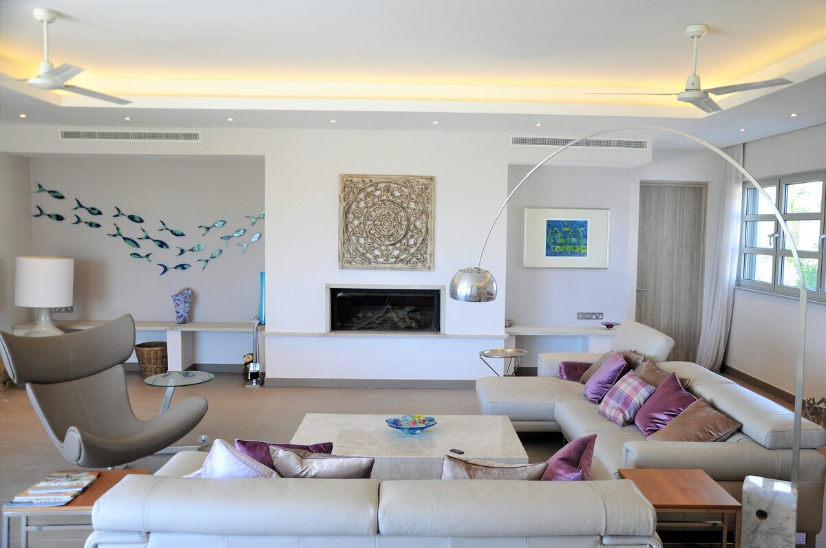 A spacious living area is shown, featuring a modern design with comfortable seating. A neutral palette is complemented by colorful cushions. A large coffee table is centered, with a decorative piece on display. Natural light floods in through multiple windows, enhancing the airy feel.