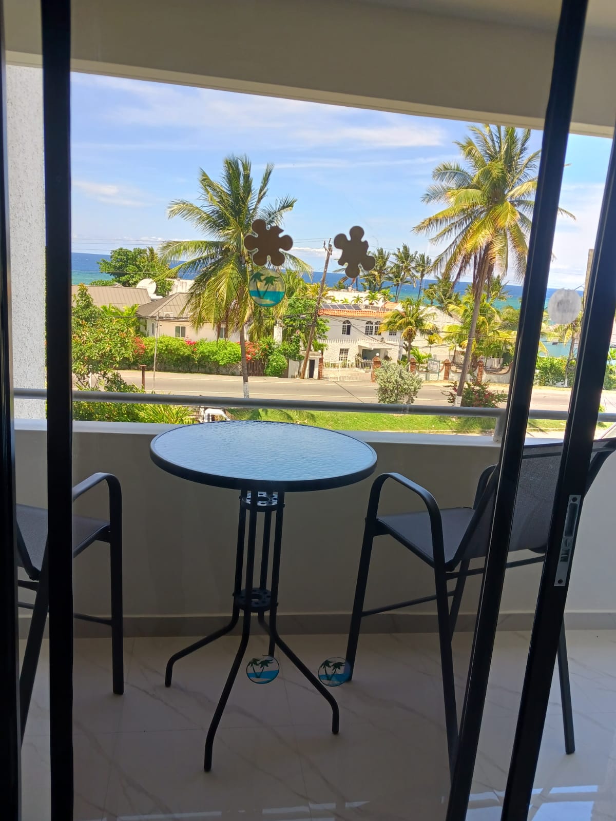 A balcony features a small round table accompanied by two chairs, offering a clear view of palm trees and nearby buildings. The ocean can be seen in the distance under a bright sky.