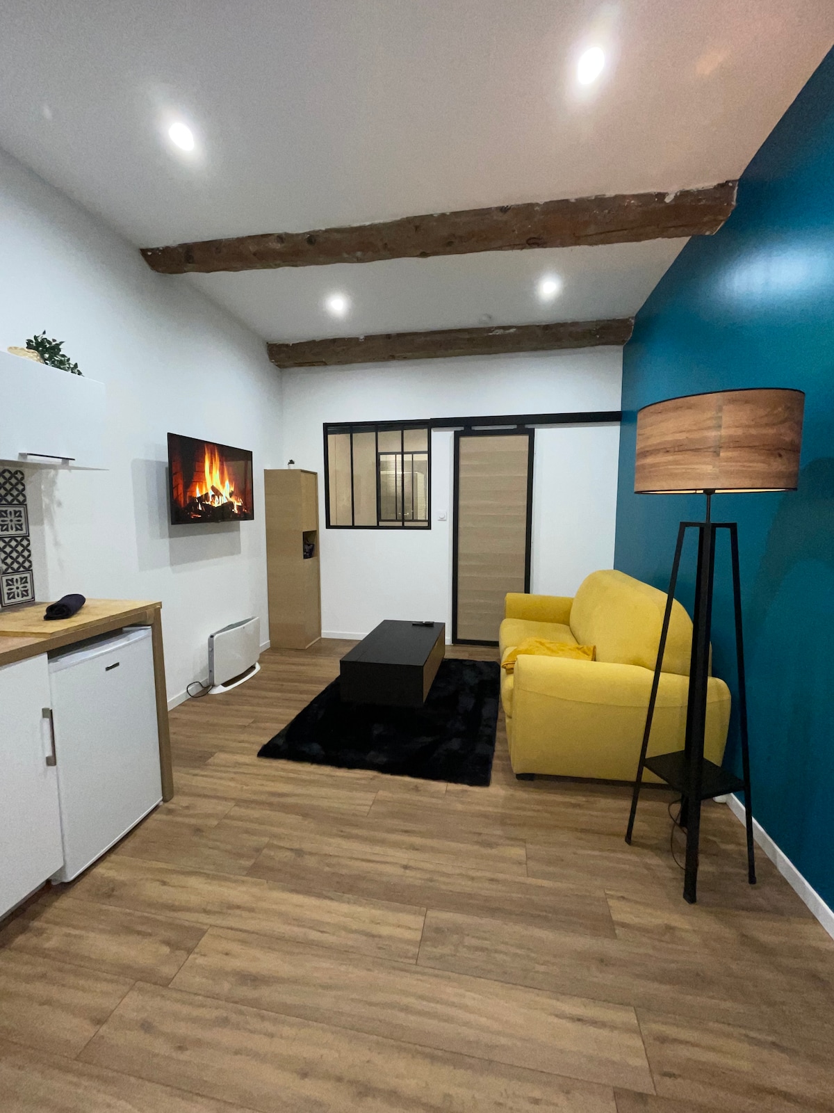 A cozy living space features a yellow sofa, a black coffee table, and a wall-mounted television displaying a virtual fireplace. Wooden beams line the ceiling, and a floor lamp stands beside the sofa. The walls are painted in a mix of white and blue, enhancing the modern decor.