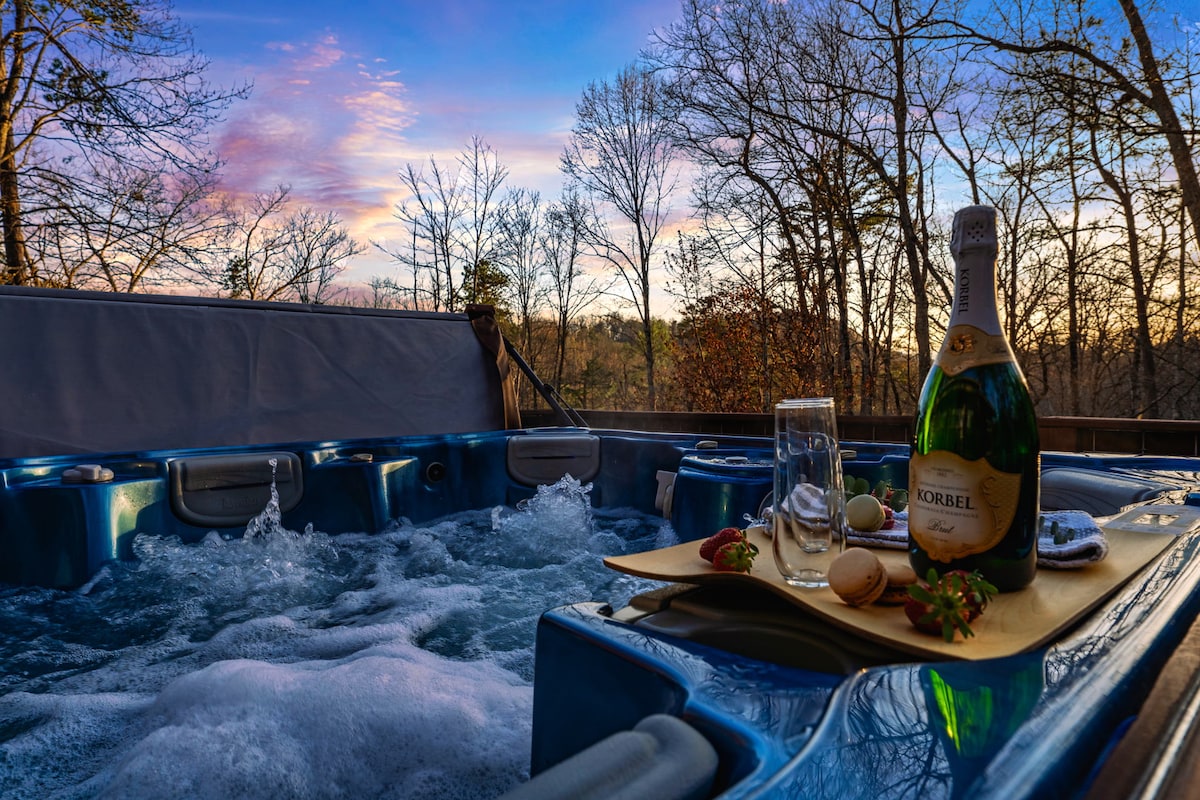 A luxury hot tub with bubbling water is shown in the foreground, complemented by a bottle of champagne and two glasses on a tray. The surrounding area features trees silhouetted against a colorful sunset sky, creating a serene outdoor relaxation space.