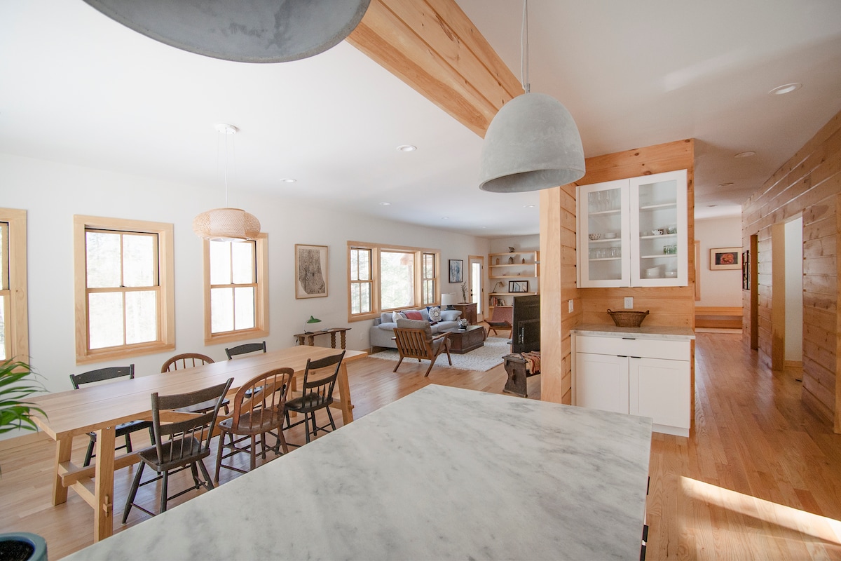 An open-concept living area showcases warm wood accents and large windows, inviting natural light. A spacious dining table is set for gatherings, while a cozy seating area is visible in the background. The modern kitchen features a marble countertop and stylish pendant lights overhead.