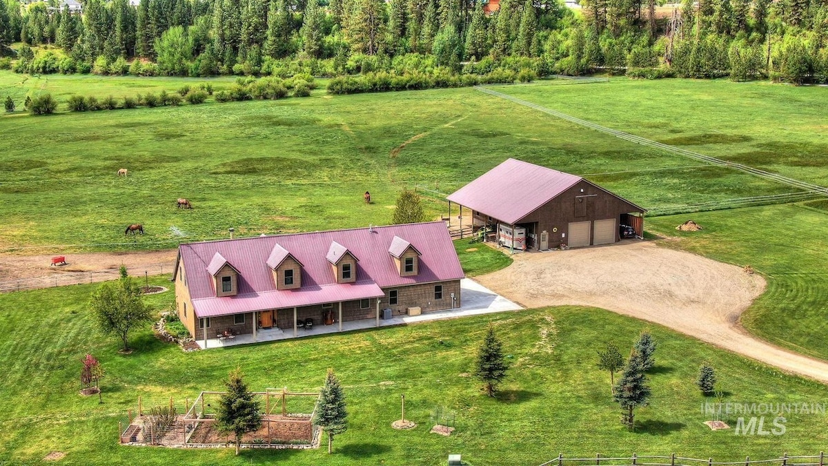An aerial view captures a spacious property set against a green landscape. A large home with a red roof is seen alongside a barn, both surrounded by grassy fields and trees. Horses can be observed grazing in the distance, enhancing the rural setting.