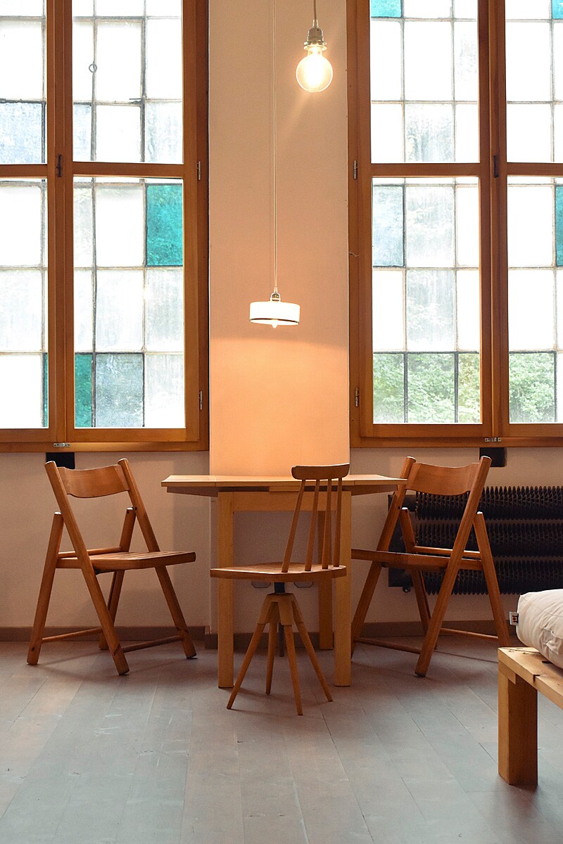 A cozy corner is created with a small wooden table supported by three classic wooden chairs. Two large windows allow natural light to fill the space, highlighting the light-colored walls and wooden flooring. A simple pendant light hangs above the table.