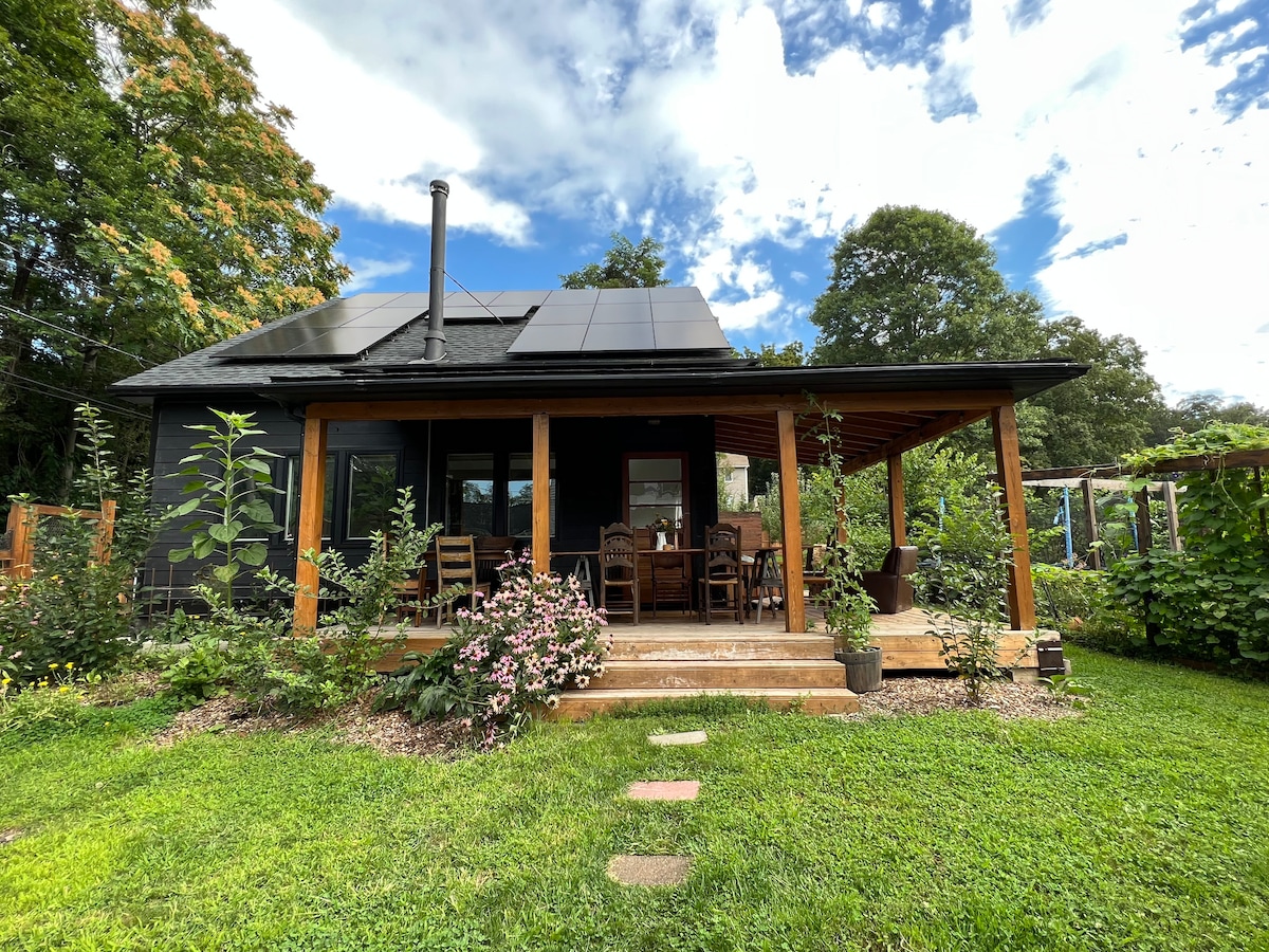 A black guesthouse features a covered deck with a dining area and seating, surrounded by gardens. Solar panels are installed on the roof, and a lush green lawn enhances the outdoor space. Tall plants and flowers add visual interest to the surroundings.