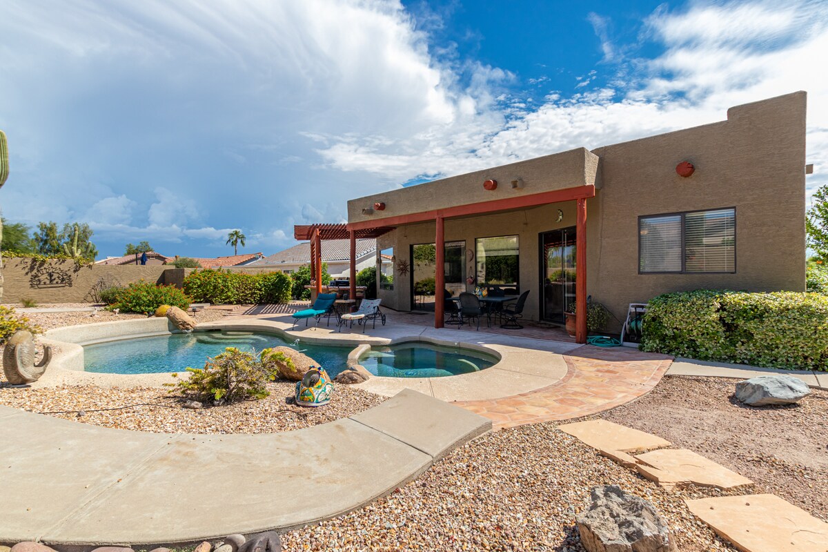 A spacious backyard features a shimmering pool and a jacuzzi surrounded by desert landscaping. Comfortable seating is arranged under a patio, providing shaded areas. The home’s exterior showcases Southwest architectural elements, with large windows offering picturesque views of the surrounding landscape.