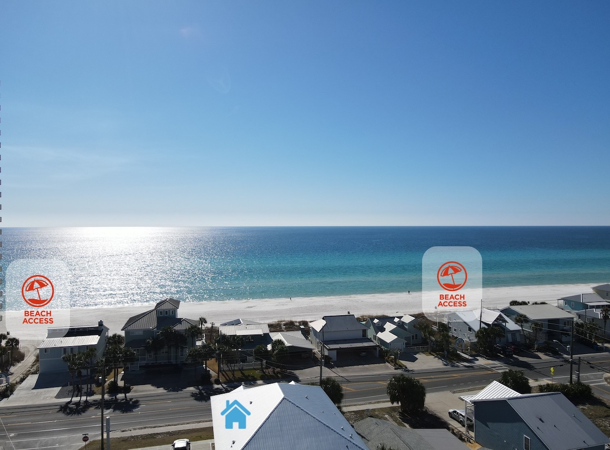 A panoramic view showcases the clear turquoise water meeting the soft white sand beach under a bright blue sky. Beach access points are marked, indicating easy access to the shoreline. Nearby structures are visible, providing context to the coastal landscape.