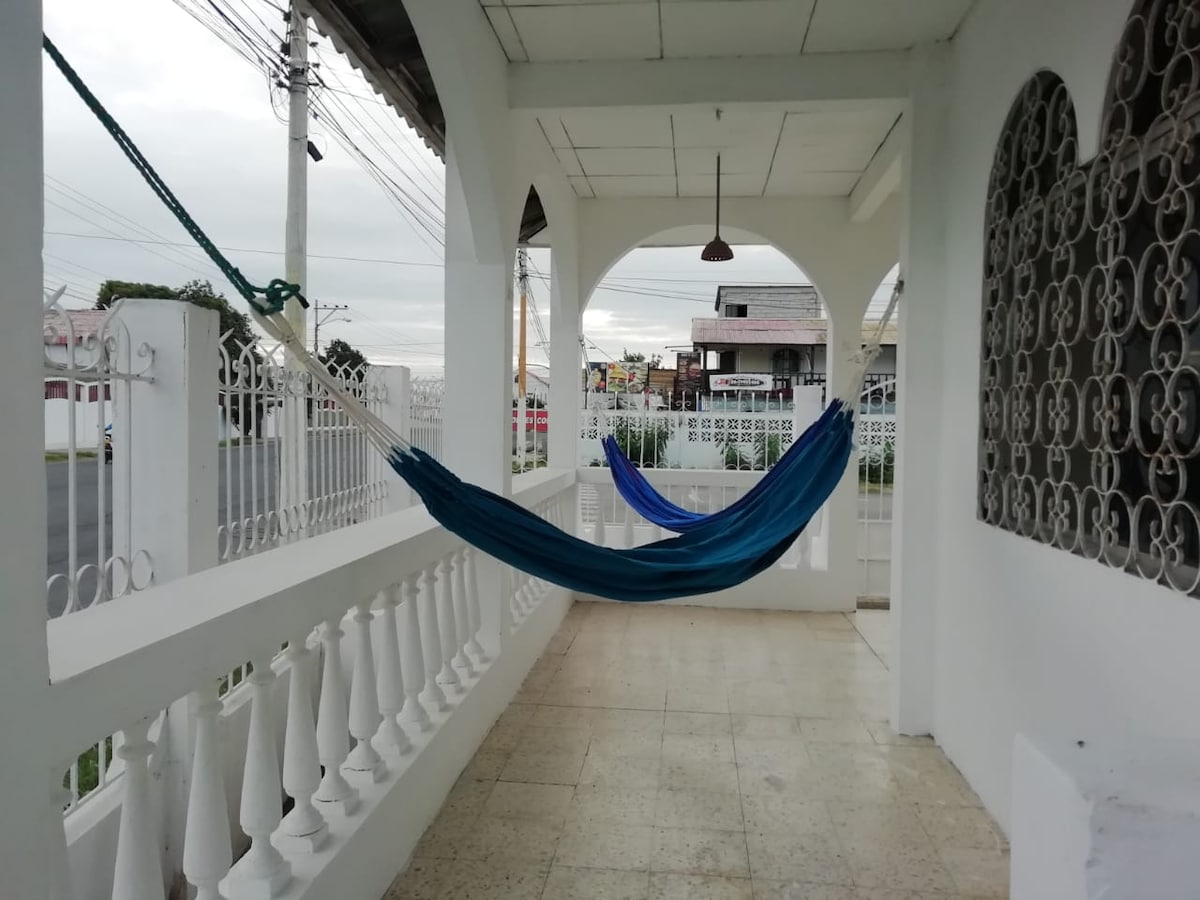 A spacious porch features two hammocks, one deep blue and the other lighter in shade, hanging between decorative columns. The area is enclosed by a white railing and adorned with intricate ironwork, allowing for a view of the street outside.
