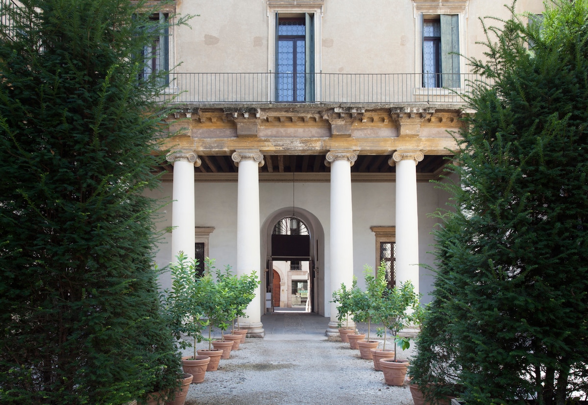 The entrance of a historic building is flanked by tall columns and lined with potted plants. A pathway leads through an arched entryway, with greenery on both sides enhancing the welcoming atmosphere. The intricate architectural details are visible, reflecting the elegance of the era.