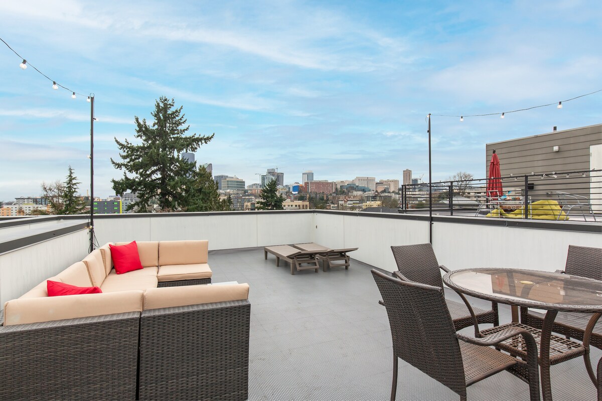 The rooftop deck features a seating area with a sectional sofa adorned with bright red cushions. A dining table with chairs is positioned nearby, offering views of the Seattle skyline against a clear blue sky.