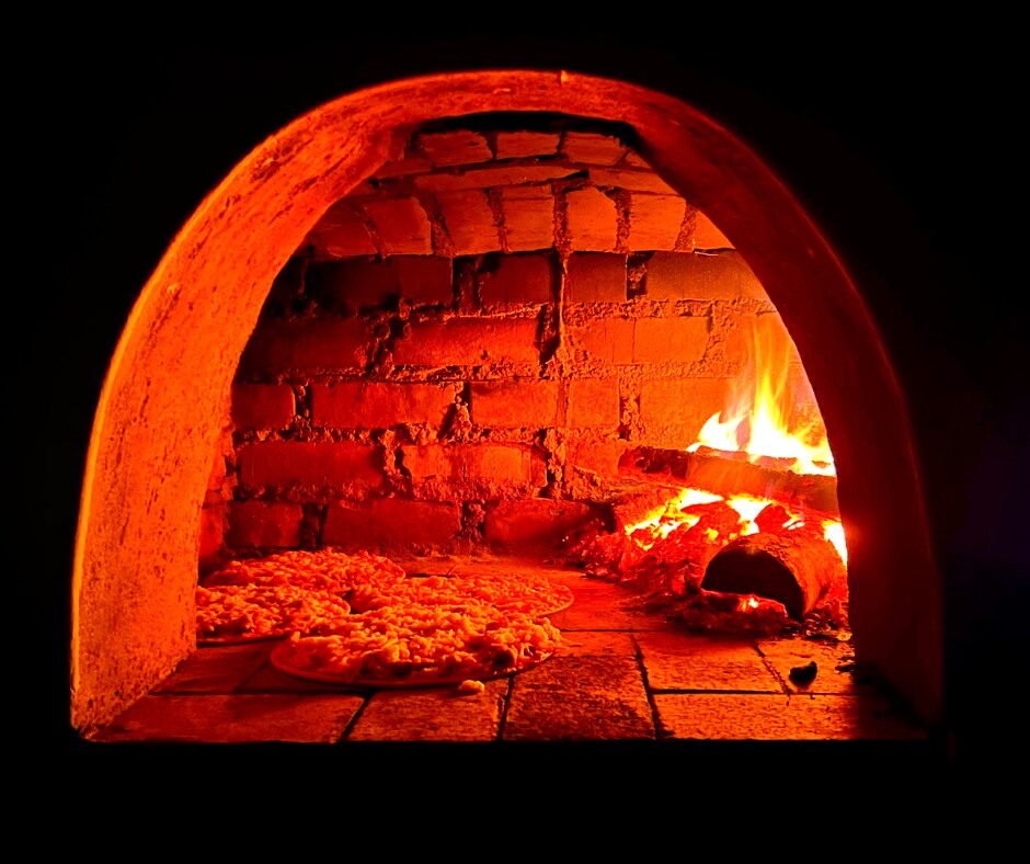 A traditional pizza oven is shown, with glowing embers and flames providing warmth. Two pizzas are visible on the stone floor inside the oven, indicating a rustic cooking experience. The brick walls of the oven suggest durability and craftsmanship.