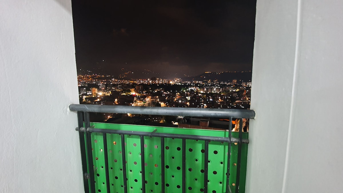 A balcony is depicted with a green railing, offering a view of the urban landscape at night. City lights twinkle in the distance, revealing the layout of buildings and the peaks of nearby hills under a dark sky.