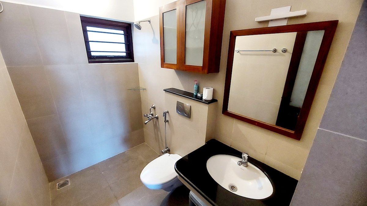 The bathroom is designed with neutral tones, featuring a wall-mounted mirror above the sink. A modern toilet is positioned beside a sleek shower area enclosed by glass. Natural light enters through a small window, enhancing the space's openness.
