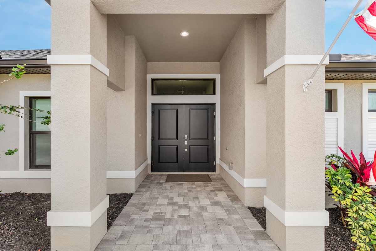 The entrance features a double door design in dark tones, framed by light-colored walls. Paved flooring leads to the doors, with garden greenery and colorful plants flanking the entrance, creating an inviting approach to the home.