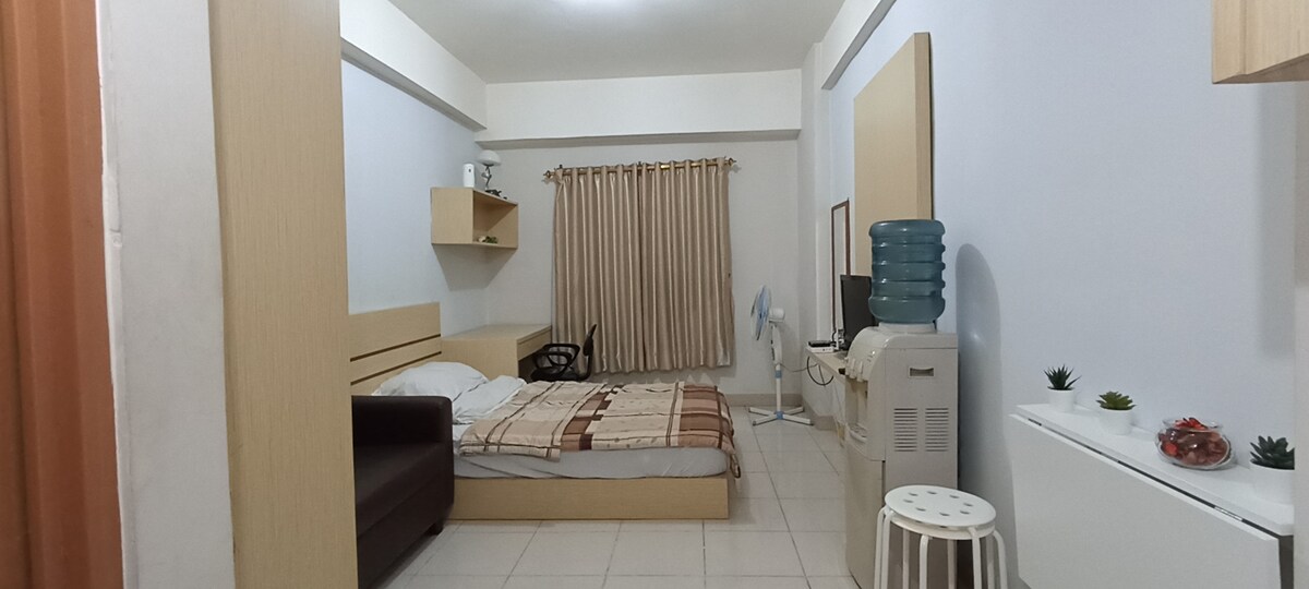 A spacious studio room is furnished with a small double bed, a sofa, and a working desk with a chair. Natural light enters through the window, while a refrigerator and a water dispenser are present. Neutral colors and clean lines create a calm environment.