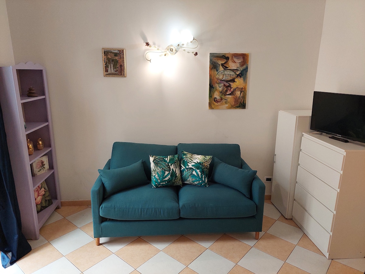 A cozy seating area features a teal sofa adorned with tropical print cushions. To the left, a purple bookshelf holds decorative items, while a white dresser and a television are positioned nearby. Light is provided by an artistic wall fixture and natural sources.