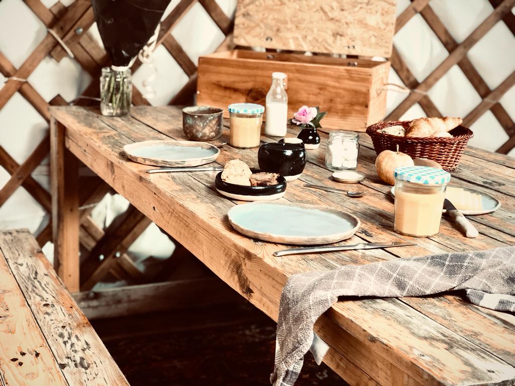 A rustic wooden dining table is set with blue plates and various jars filled with breakfast items. A basket of pastries rests alongside a small jar of cream and a bottle of milk. A towel is draped casually over the table's edge, adding to the natural charm.