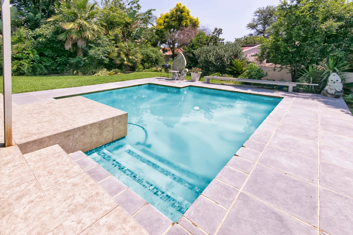 A serene pool area is showcased, surrounded by lush greenery. The pool features a rectangular shape with a shallow entry, framed by textured stone tiles. A small seating area is visible beside the pool, providing a calm spot to relax.