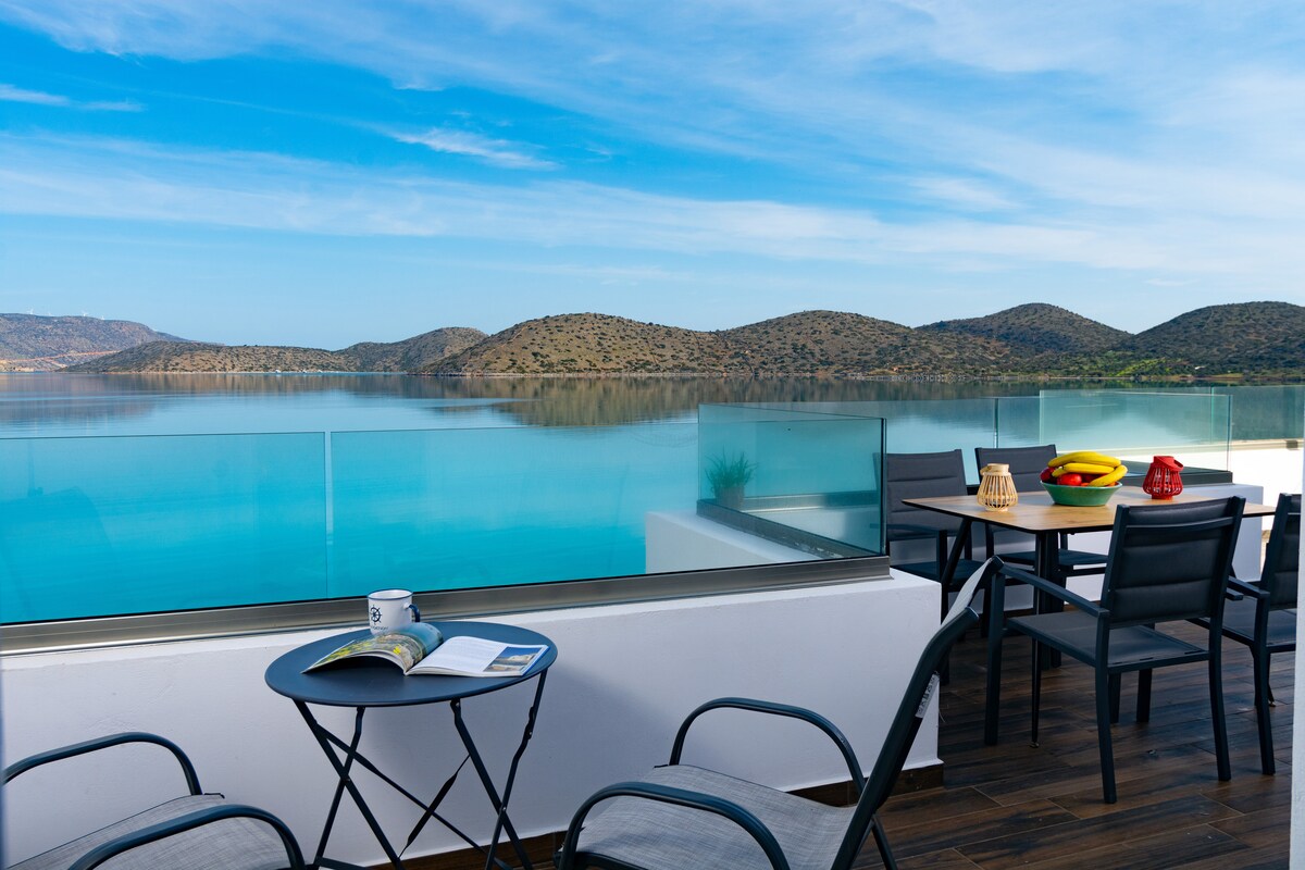 A balcony is shown with a table set for dining, accompanied by chairs. A serene view of the turquoise water extends toward distant hills under a clear sky. A small round table holds a book and a coffee mug, enhancing the space's tranquility.