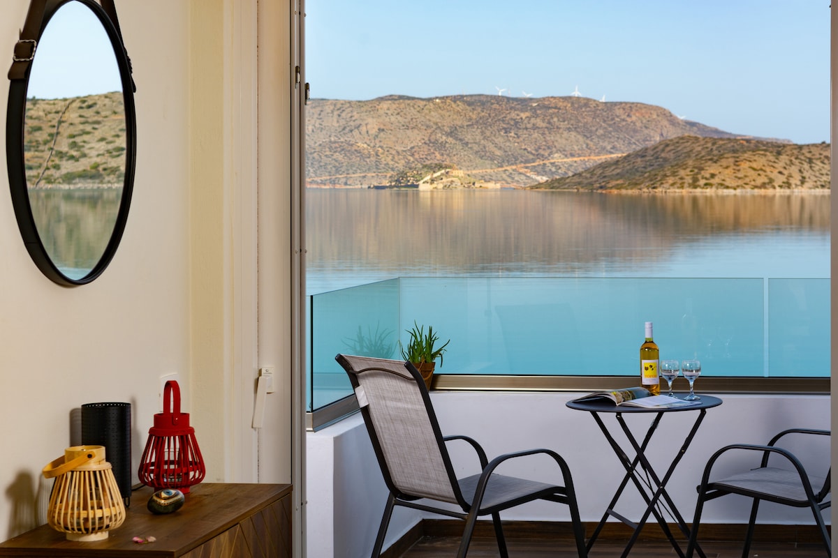A small balcony features a round table with two chairs, adjacent to a large window offering views of the calm sea and distant hills. A bottle of wine and two glasses are placed on the table, complemented by decorative items on a nearby sideboard.