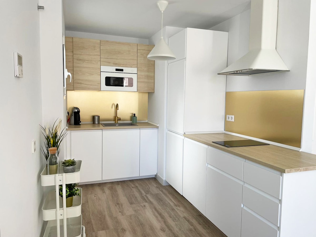 A modern kitchen is displayed featuring a blend of light wood and white cabinetry. Stainless steel appliances, including an oven and refrigerator, are integrated. A sleek countertop extends alongside a minimalist sink, while a small plant adds a touch of greenery to the space.