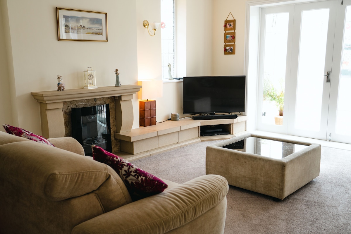 A comfortable lounge area features a light-colored sofa adorned with decorative pillows. A modern coffee table sits centrally on soft carpet. A television is positioned against the wall, and large glass doors allow natural light to fill the room, leading to a small outdoor space.