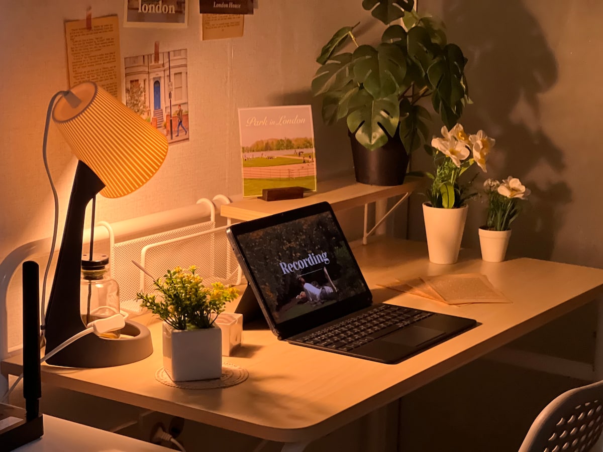 A well-lit workspace is presented with a wooden desk, featuring a laptop displaying the word 'Recording.' A stylish lamp provides soft lighting, while potted plants add a touch of greenery. Elegant floral arrangements enhance the atmosphere, offering a serene and organized environment.