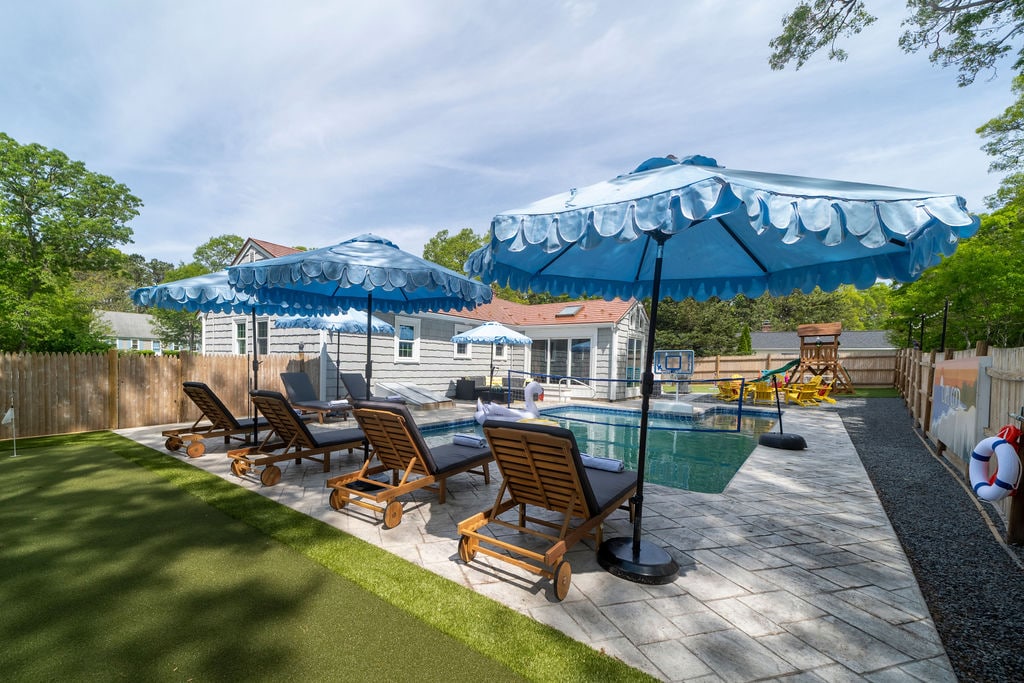 A sunny outdoor area is showcased, featuring a private heated pool surrounded by lounge chairs. Large blue umbrellas provide shade next to the pool, while decorative landscaping and a wooden playset are visible in the background. The space invites relaxation and comfort.