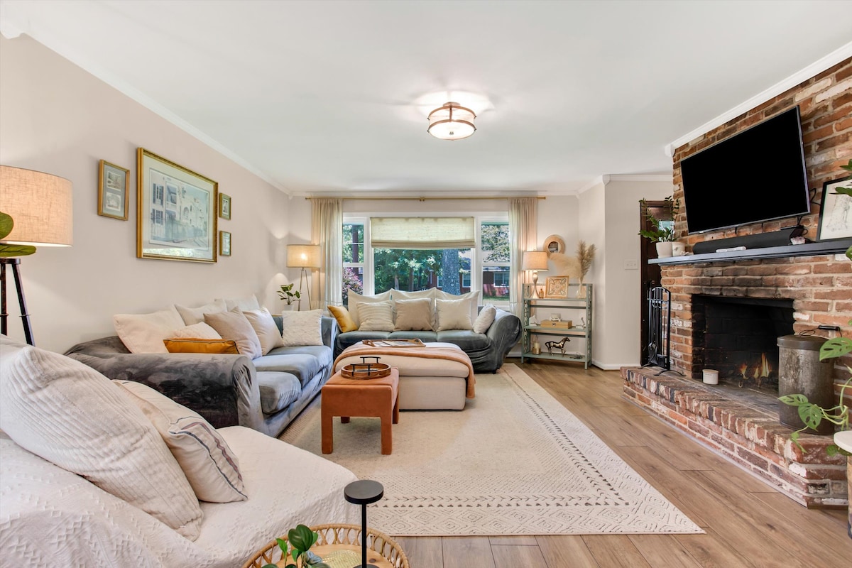 A comfortable living area showcases a mix of light and dark furnishings, including a large sectional sofa and an accent chair. A soft area rug is positioned on the floor, and natural light streams through the window. A stone fireplace with a mounted television adds character to the space.