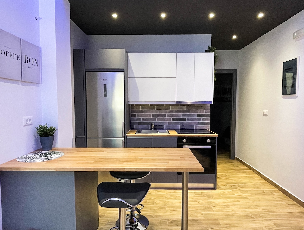A modern kitchen space is presented with sleek cabinetry and stainless steel appliances. A wooden bar counter with a black stool features a minimalistic design. The backsplash is adorned with gray tiles, enhancing the contemporary aesthetic of the room, which is illuminated by recessed lights.