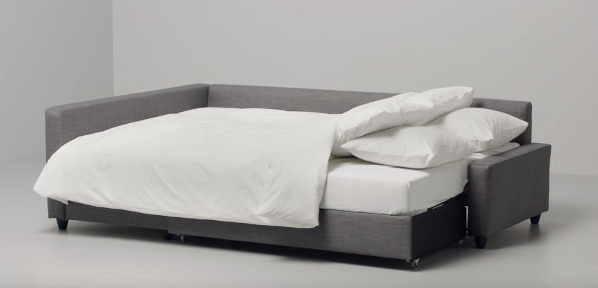 A modern sofa bed is displayed in an open layout, featuring a sleek gray fabric. The bedding is neatly arranged, with a white duvet and fluffy pillows resting on a spacious mattress, illustrating a comfortable sleeping arrangement.