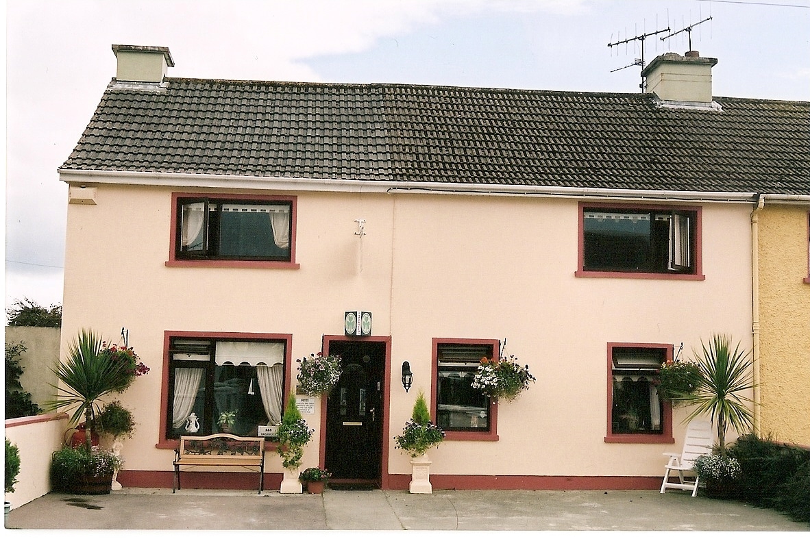 A two-story home is depicted with a peach-colored exterior and decorative red trim around windows and doors. Potted plants adorn the windowsills, and a small seating area is visible on the right. The roof is covered with gray tiles, and an antenna is situated above.