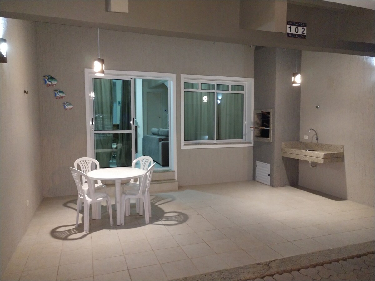 An outdoor patio features a round dining table surrounded by four white chairs. Sliding glass doors lead into a cozy indoor space, while a large sink and counter are positioned along the wall. Soft lighting provides illumination.
