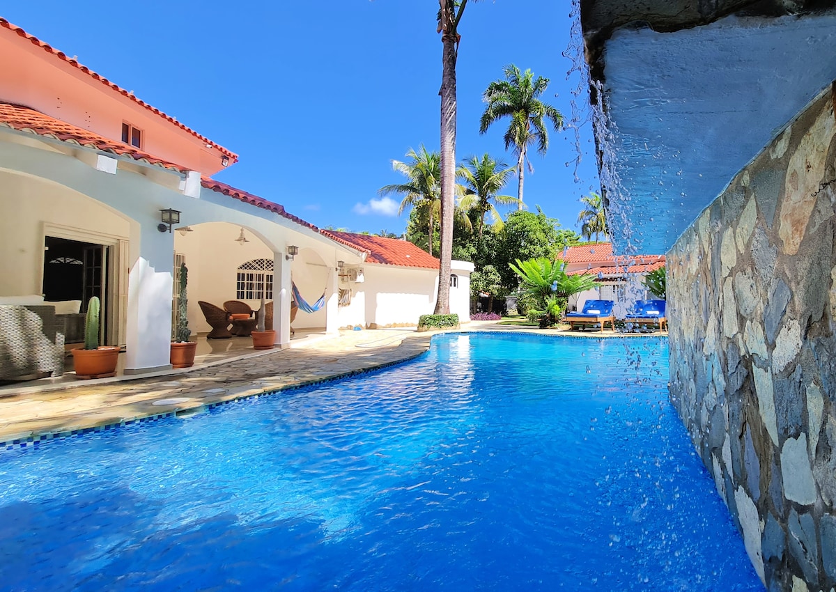A large free-form swimming pool is highlighted, with a cascade gently flowing from a stone wall. Surrounding palm trees and tropical gardens add to the serene setting. The villa's exterior, featuring casual seating areas and a hammock, is visible in the background.