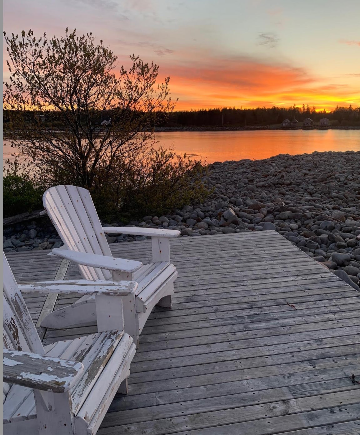 A wooden deck features two weathered white chairs positioned to overlook a serene waterfront at sunset. The sky is painted with hues of orange and pink, reflecting on the calm water, while smooth stones border the shore, complementing the tranquil scene.