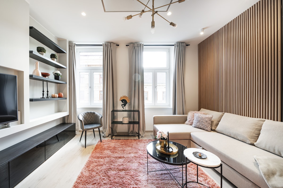 A modern living area features a comfortable sectional sofa and a plush area rug. Natural light stream through large windows, complemented by elegant curtains. Shelving units and decorative items enhance the space, while a stylish chair adds seating. A circular coffee table holds decorative accessories.