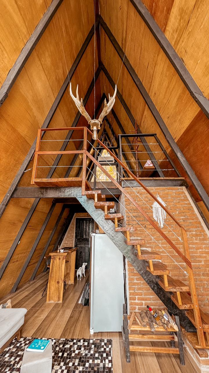 An interior view of a cabin features a warm wooden decor with high ceilings. A staircase made of wood leads to a loft area, complemented by brick walls. Unique antlers are displayed overhead, and natural light streams through windows, casting soft shadows on the floor.
