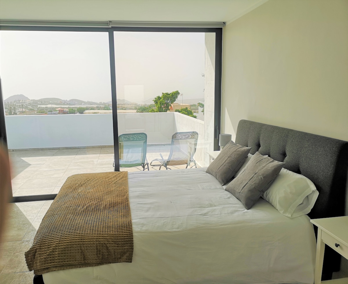 A spacious bedroom features a comfortable bed adorned with neutral linens and decorative pillows. Large glass doors provide a clear view of an outdoor terrace furnished with two chairs, allowing for natural light to fill the room while offering expansive views of the surrounding landscape.